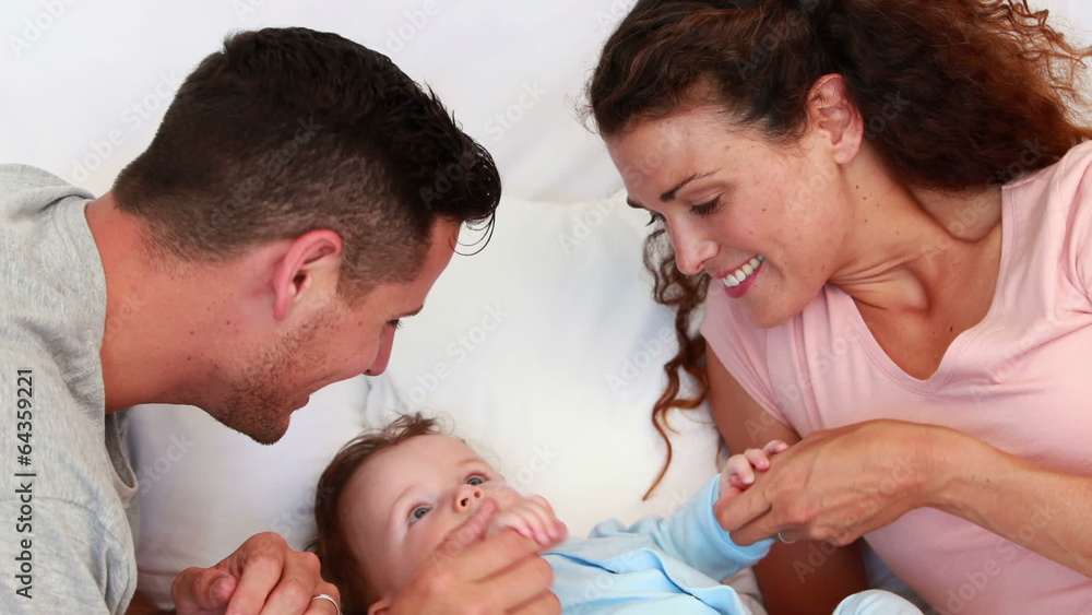 Baby boy in blue babygro being tickled by happy parents on bed Baby boy in blue babygro being tickled by happy parents on bed