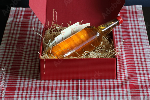 Red box filled with straw, bottle and european money, standing on a checked table-cloth.  photo