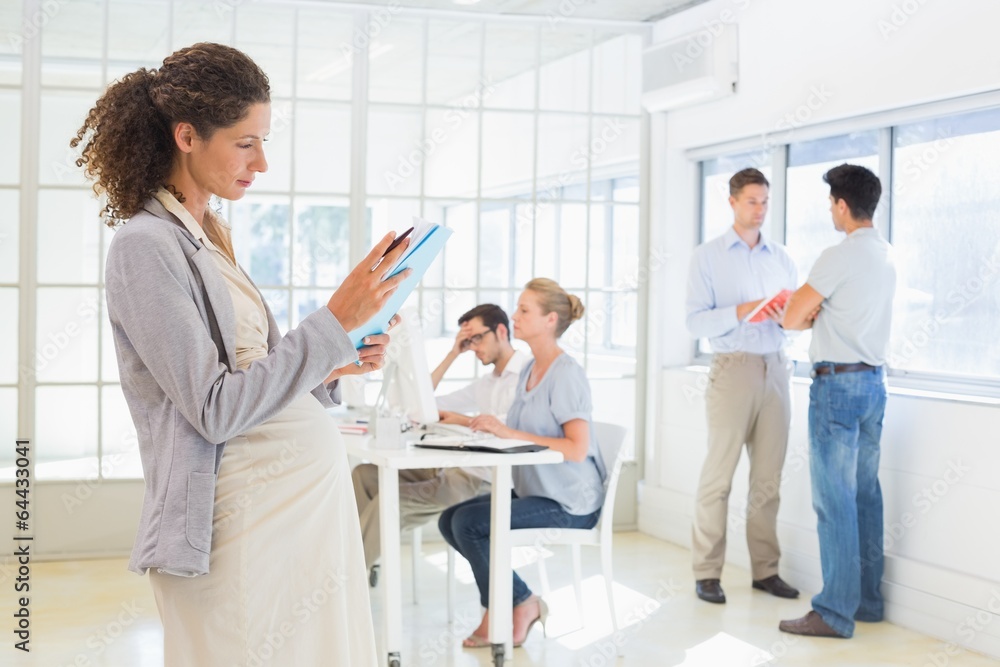Pregnant businesswoman reading file with team behind her Pregnant businesswoman reading file with team behind her