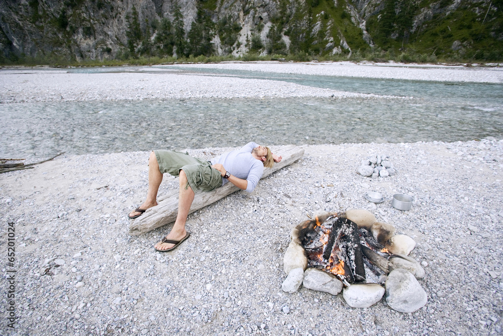 Fototapeta premium Deutschland,Bayern,Tölzer Land,Junger Mann auf Baumstamm in der Nähe von Lagerfeuer ruhen