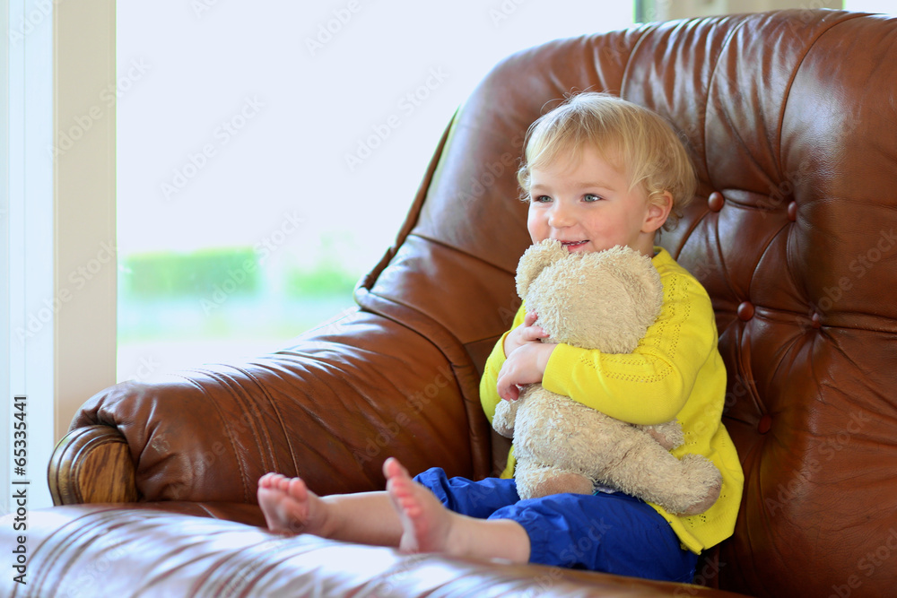 Fototapeta premium Cute toddler girl sitting on sofa playing with teddy bear