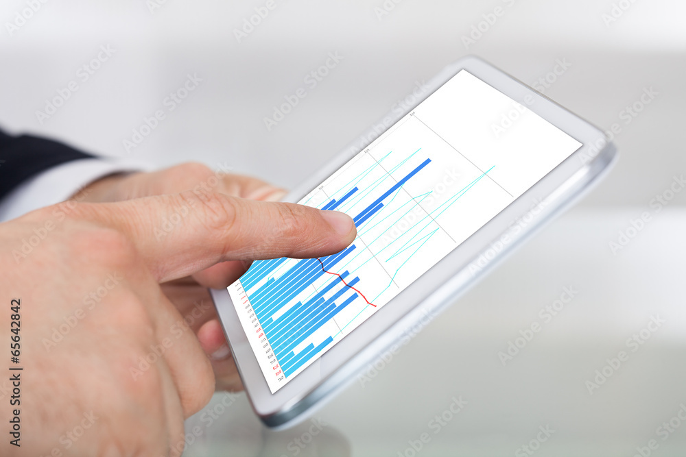 Businessman Comparing Graphs On Digital Tablet At Office Desk