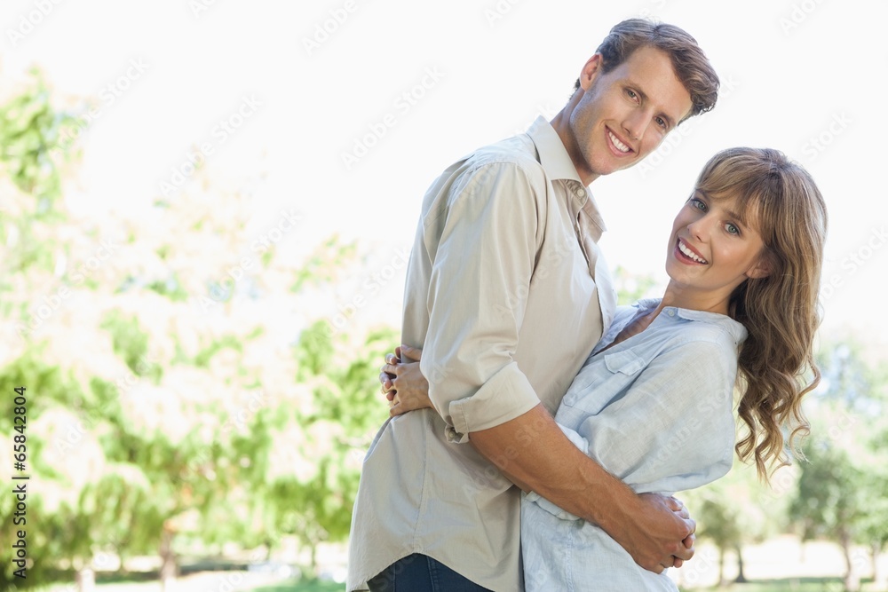 Carefree couple standing in the park and hugging smiling at came Carefree couple standing in the park and hugging smiling at came