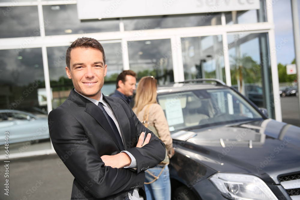Smiling car dealer standing by vehicle, clients in background Smiling car dealer standing by vehicle, clients in background