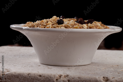 Side view of bowl of homemade granola with oats, almonds, organic raisins, and walnuts on a black background and travertine tile