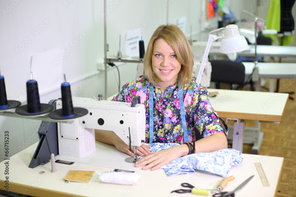 Pretty female tailor sits at table with sewing machine, sews Pretty female tailor sits at table with sewing machine, sews