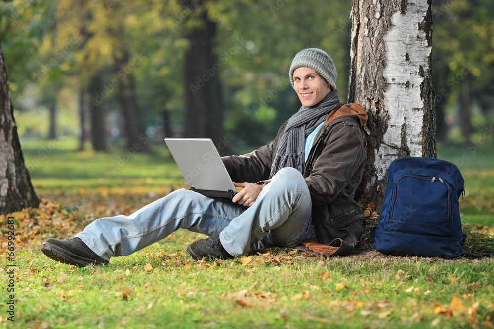 Young man working on laptop seated by a tree in park Young man working on laptop seated by a tree in park