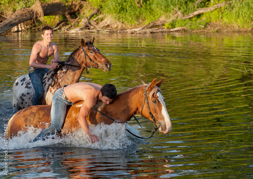 Two young men astride horses go down the river