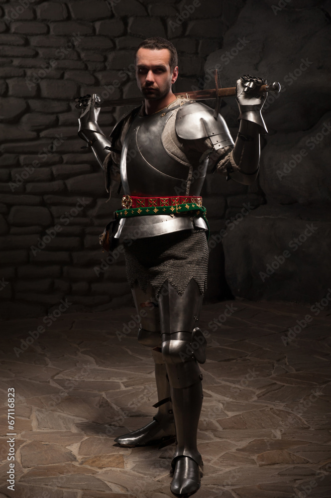 Fototapeta premium Medieval Knight posing with sword in a dark stone background