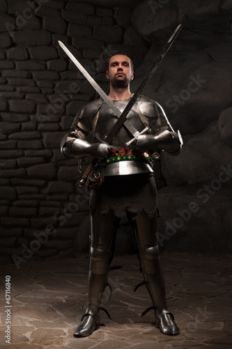 Medieval Knight posing with two swords on in a dark stone wall b