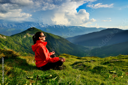 Young woman in red jacket sitting in yoga pose in mountains