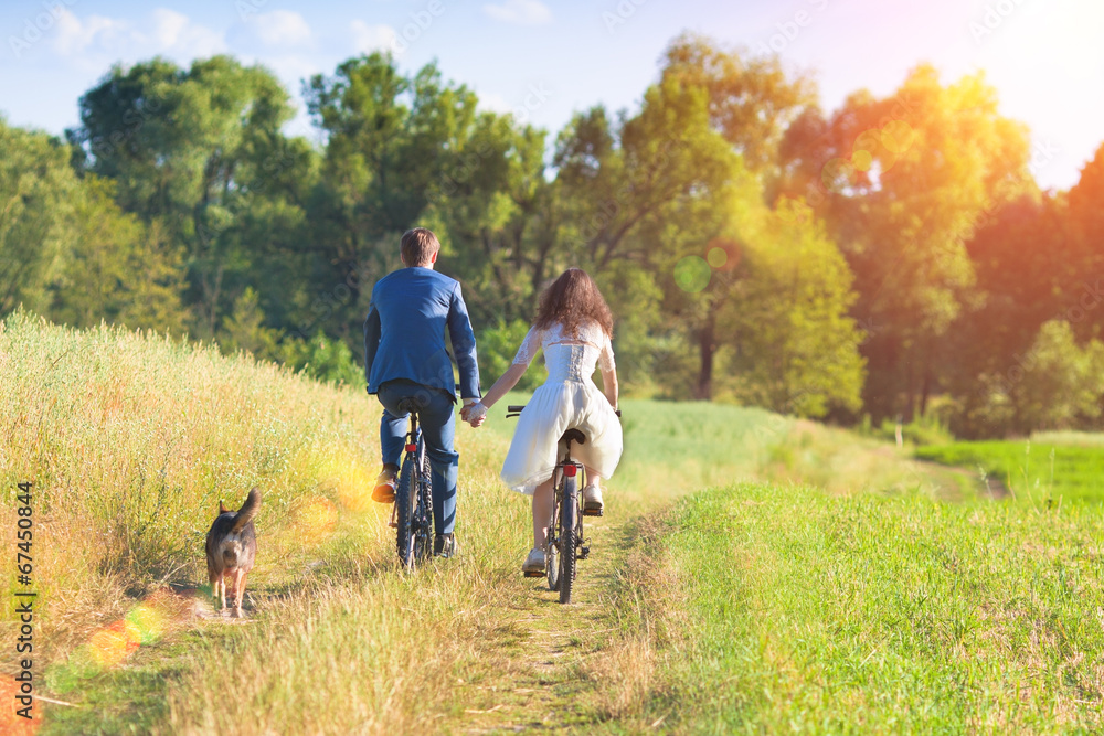 Fototapeta premium Young happy bride and groom ride bicycles in the meadow