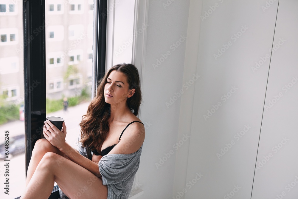 Young lady relaxing on window sill with a cup of coffee Young lady relaxing on window sill with a cup of coffee