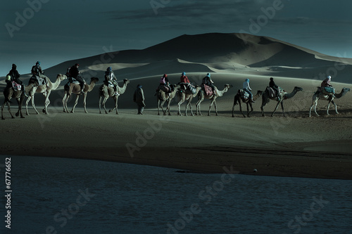Caravan of tourists passing desert lake on camels in moonlight