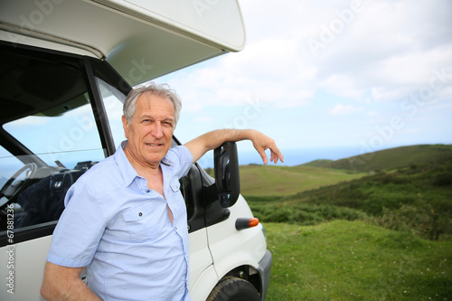 Senior man standing by camper, scenery in background