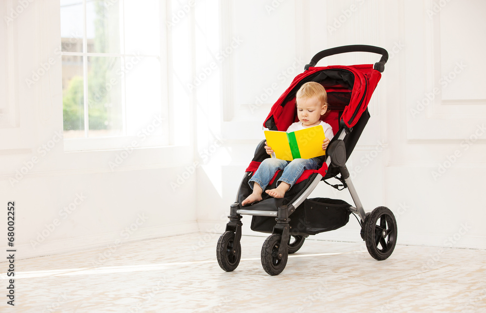 Toddler boy sitting in stroller at home and looking at open book Toddler boy sitting in stroller at home and looking at open book
