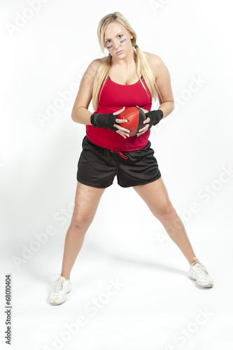 A female blond model holding a red and black football wearing a red t-shirt with black shorts on a white background.