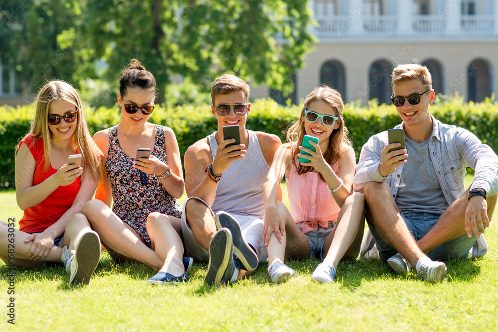 Fototapeta premium smiling friends with smartphones sitting on grass