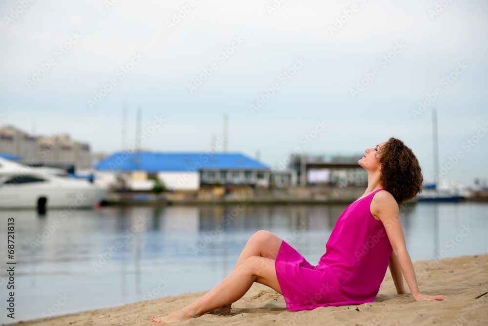 Young woman in a red dress ans curly hair sit on the water coast