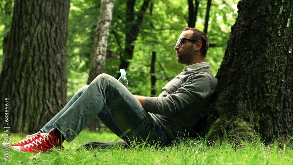 Young man drinking water while sitting by the tree in park Young man drinking water while sitting by the tree in park