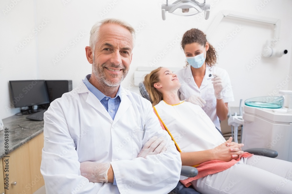 Obraz premium Dentist smiling at camera with assistant and patient behind