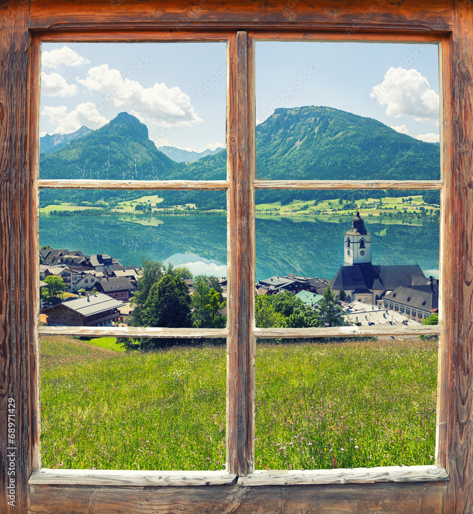Fototapeta Zamknięte stare, drewniane okno z widokiem na łąkę, dachy domów i góry w oddali
