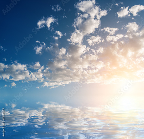 Sky with clouds and sun reflected in water surface.