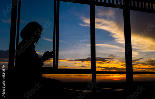 Girl playing smart phone by window at sunset with twilight sky