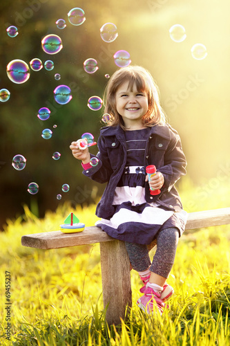 Portrait of a girl sitting on a wooden bench blows bubbles in th