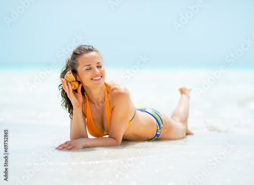 Woman laying on sea shore and listening sound of sea in shell