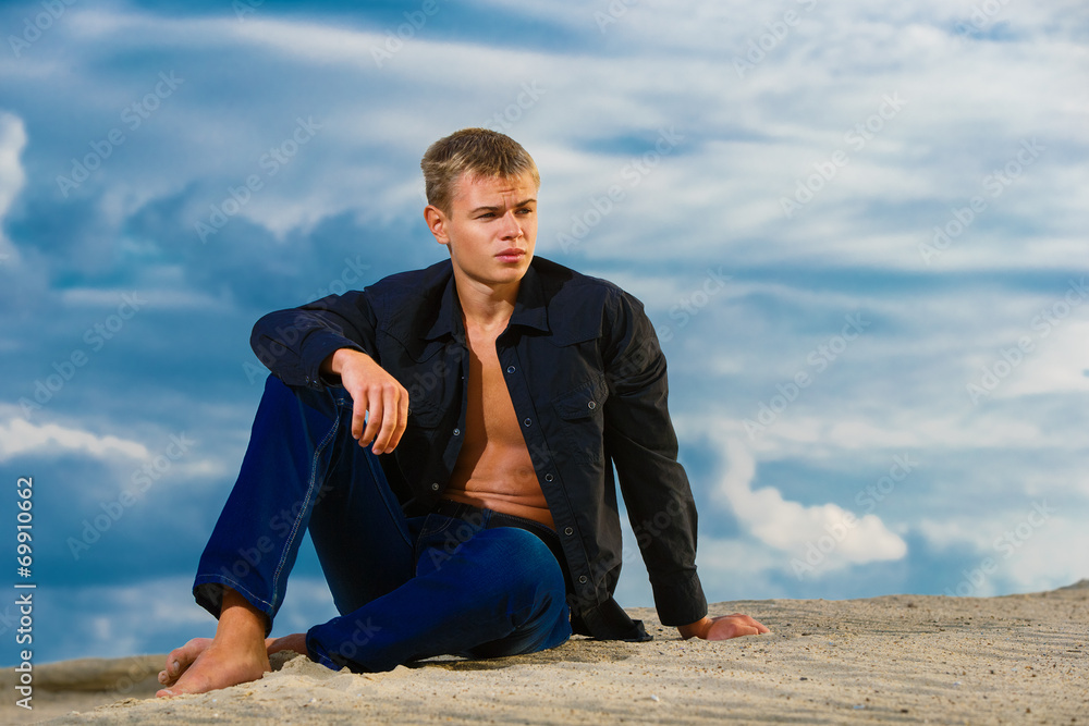 Young handsome man is sitting on the sand on the beach. Looks th