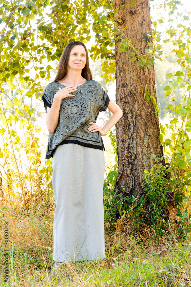 An elegant mature woman with a long dress in the shadow of a nice poplar tree in the park during a warm summer day An elegant mature woman with a long dress in the shadow of a nice poplar tree in the park during a warm summer day