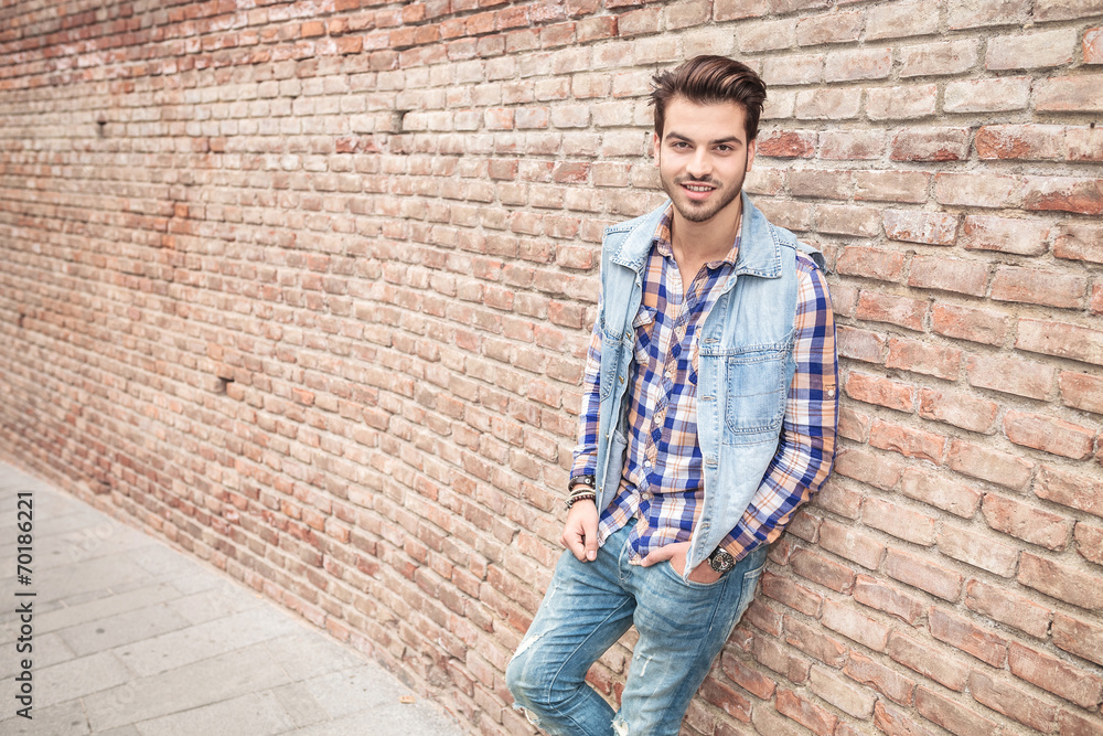young casual man smilling, leaning on a brick wall