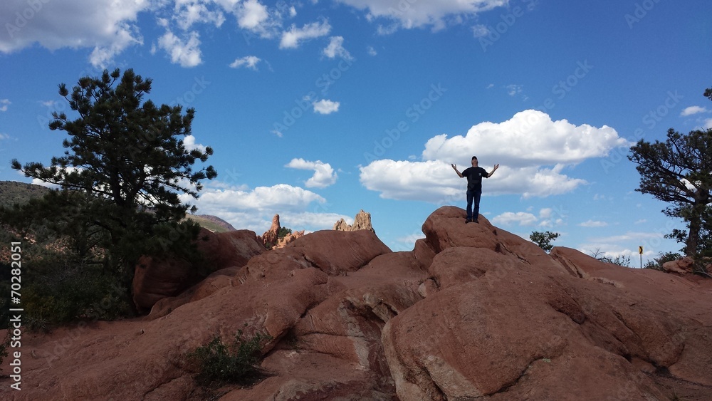 Man standing up on red rock formations in Garden of the Gods,  arms raised, holding up the clouds