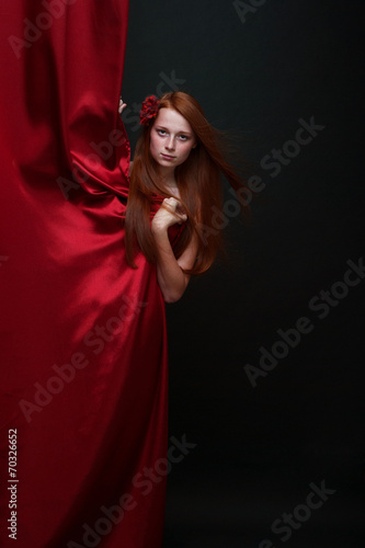 Beautiful red-haired girl looks out from behind a red theater cu © belinka