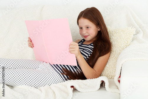 Beautiful little girl reading book on sofa in room