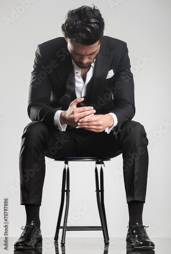 Photography man in tuxedo looking down while sitting on a stool,