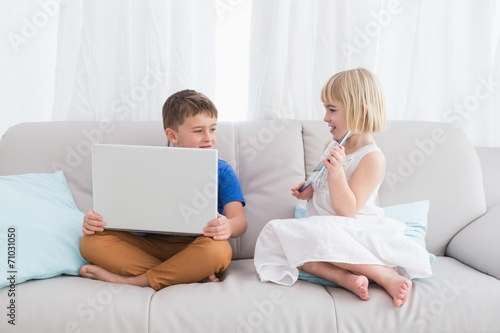 Siblings using a laptop and a tablet sitting on a couch