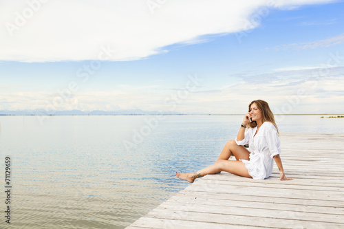 Smiling beautiful young woman sitting on a pier and talking on t