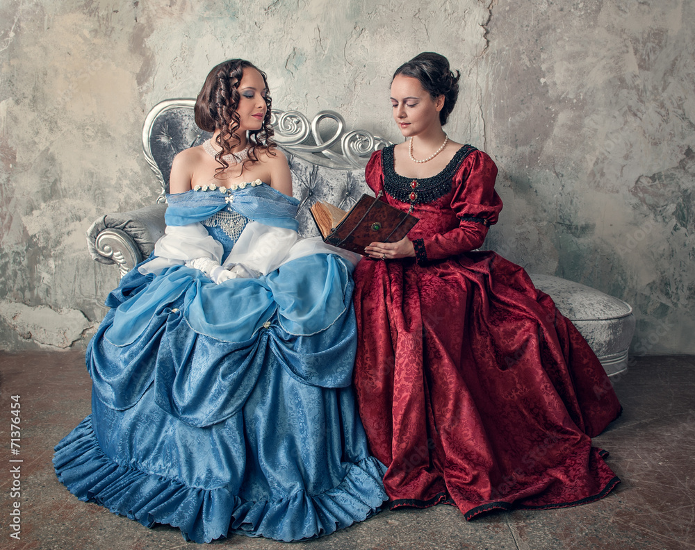 Naklejka premium Two beautiful women in medieval dresses on the sofa reading book