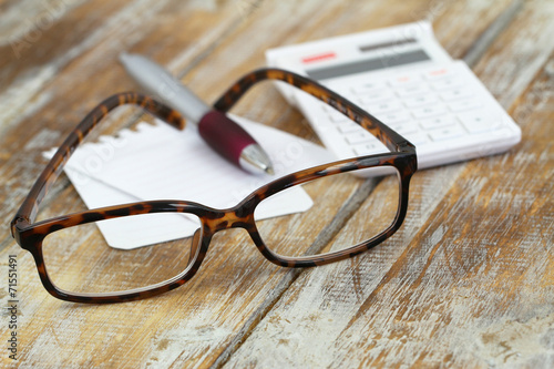Reading glasses, calculator, pen and note paper on wood