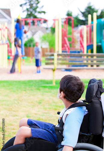 Disabled little boy in wheelchair watching children play on play