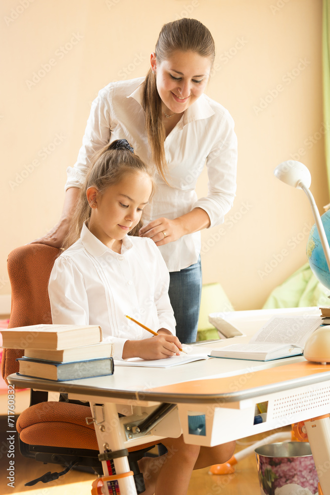 smiling woman looking at girl doing homework at bedroom