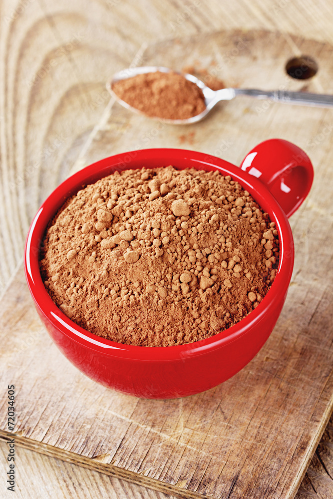 Cocoa powder in a red cup with a spoon on a rustic wooden table Cocoa powder in a red cup with a spoon on a rustic wooden table