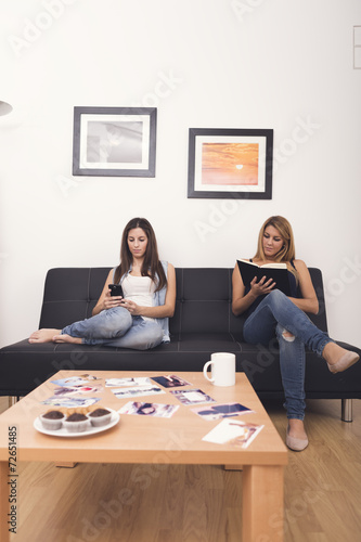 two women using mobile and reading a book on sofa in her home