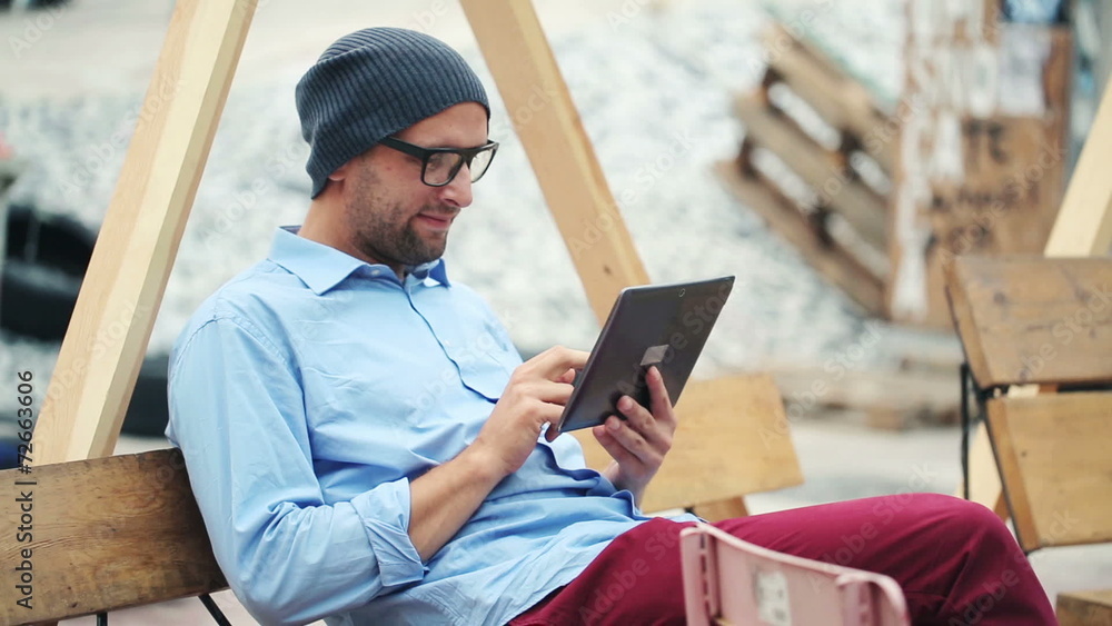 Hipster man using tablet computer while sitting on bench in the Hipster man using tablet computer while sitting on bench in the