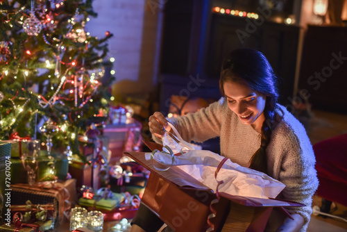 Beautiful young woman opening a gift in front of the Christmas t