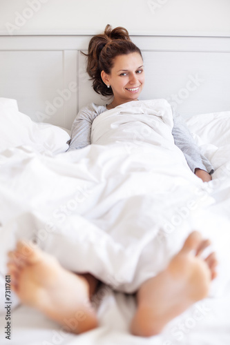 Smiling woman sits in the bed covered with soft blanket .