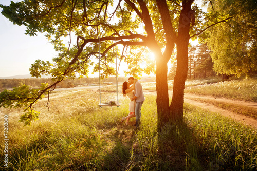Young kissing couple under big tree with swing at sunset