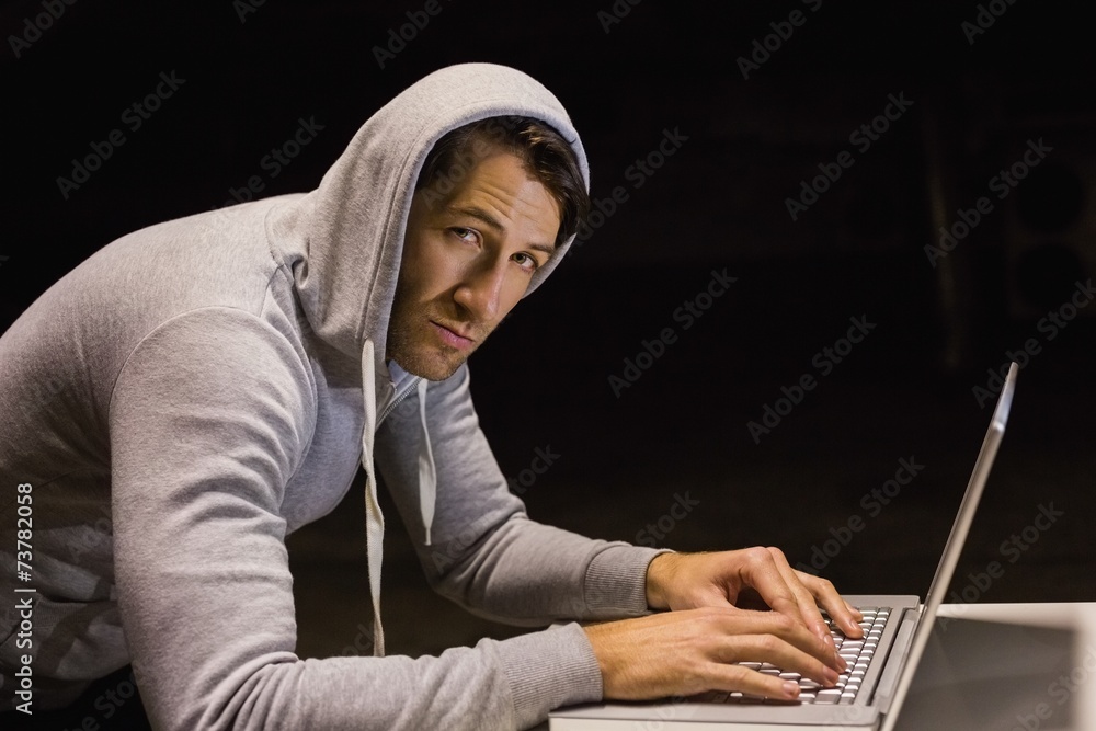 Man in hood jacket hacking a laptop while looking at camera Man in hood jacket hacking a laptop while looking at camera